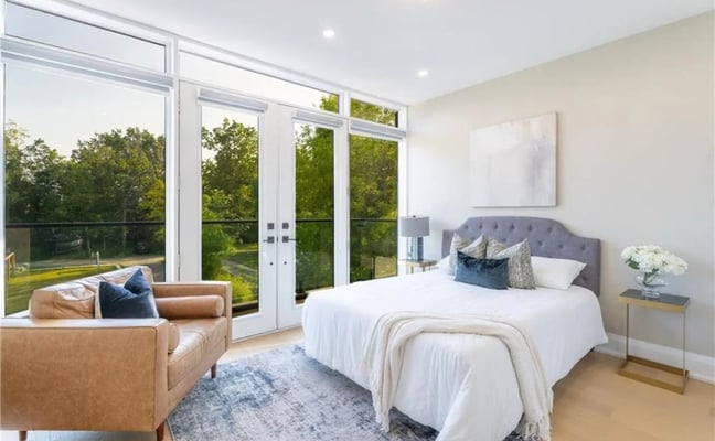 Bright bedroom with queen bed, gray upholstered headboard, tan leather chair, and floor-to-ceiling glass doors opening to green lawn.